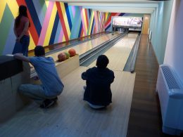 Egy tag gurítja a bowling-golyót, másik kettő pedig a pályához közel ülve nézi